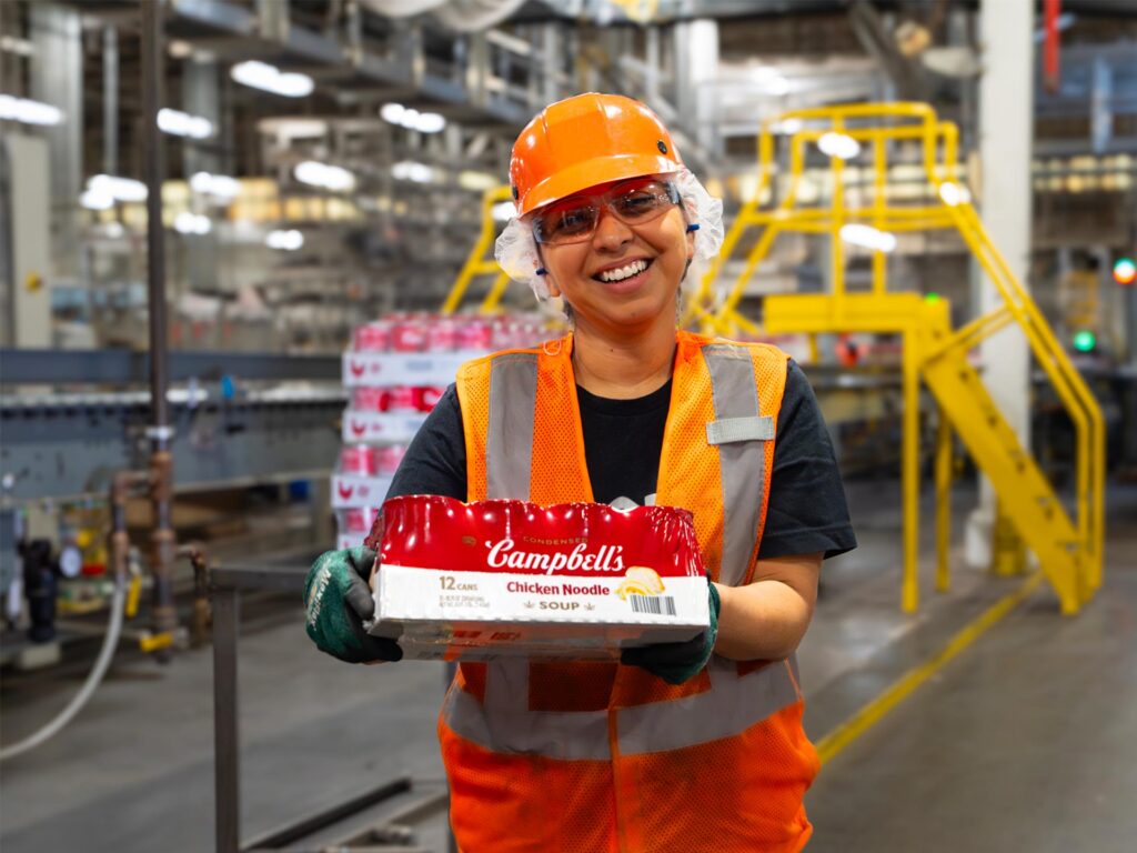 Employee holding a package of Campbell's Tomato Soup at a supply chain facility.